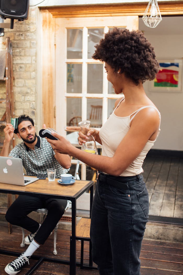 A woman processes a payment using a wireless terminal in a café setting with a customer seated nearby.